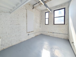 A brightly lit space with 2 large windows letting sun in against white-painted brick walls with a utility loft seen just overhead to the left