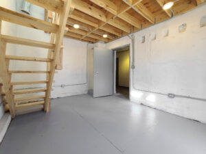 A brightly sunlit work space with white painted drywall and cement wall with a wooden staircase leading up to a second overhead work area