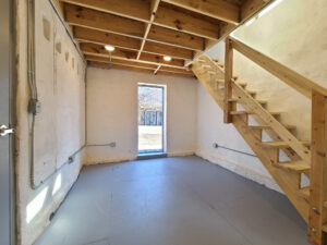 A white space with an open door leading outside from an area with bright white walls and a wooden stair case leading up a second work area above