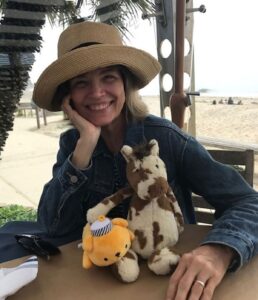 A woman with grey-blonde, neck-length hair and wearing a jean jacket and summer hat is sitting beachside at a table with two stuffed animals on a warm, sunny day