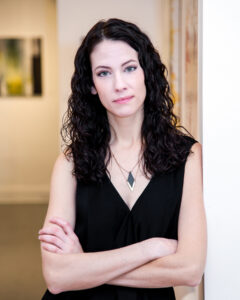 A woman with dark, wavy shoulder-length hair and wearing a black dress and a diamond shaped pendant necklace is seen in an art gallery with arms crossed looking straight ahead toward the camera