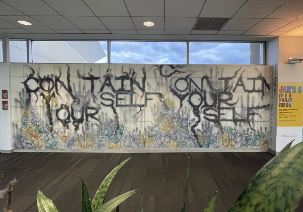 A large installation spanning an entire wall contains tiles with various purple, yellow and black lettering, black handprints and with the text "Contain Yourself" written repeatedly and more visibly in black, whispy text