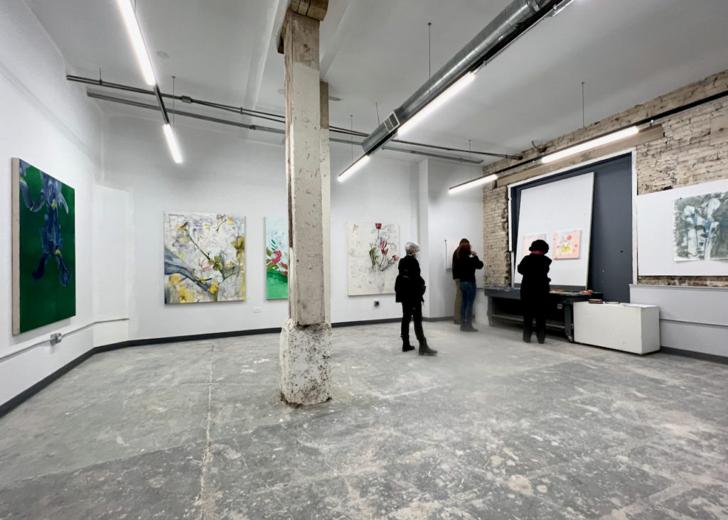 Brightly-lit studio exhibition with large work on walls and viewers walking through