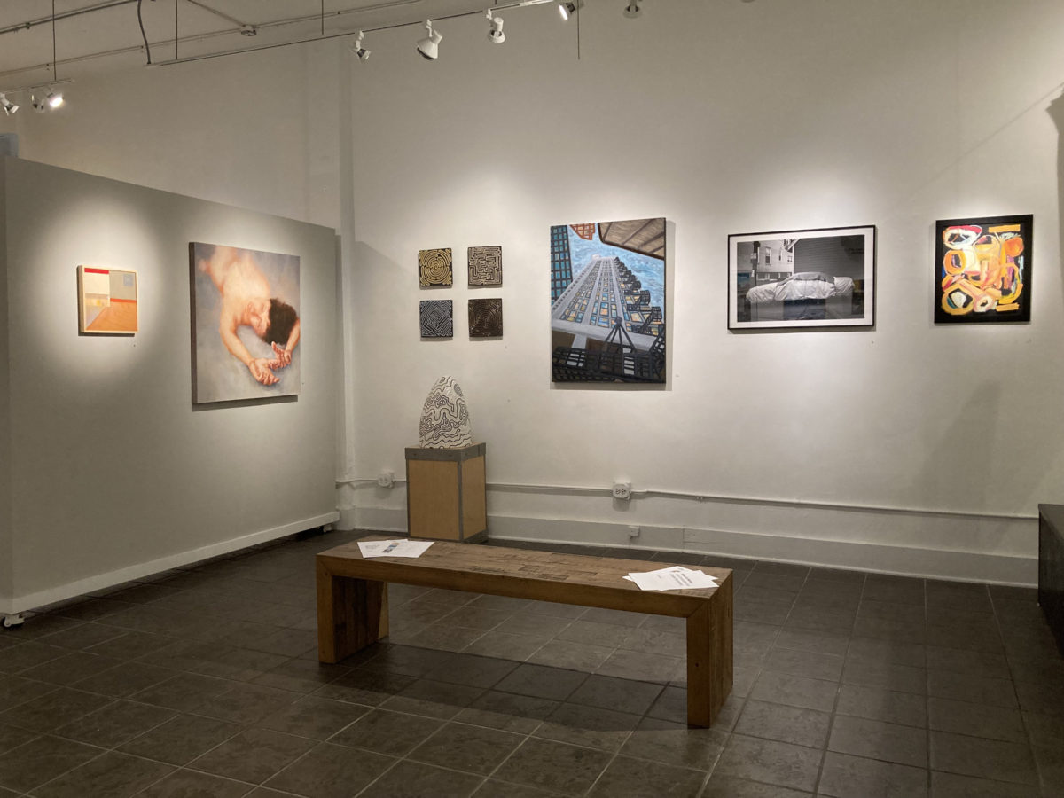 Art gallery interior with several smaller paintings along two separate walls. A small bench with show materials sits in the foreground.