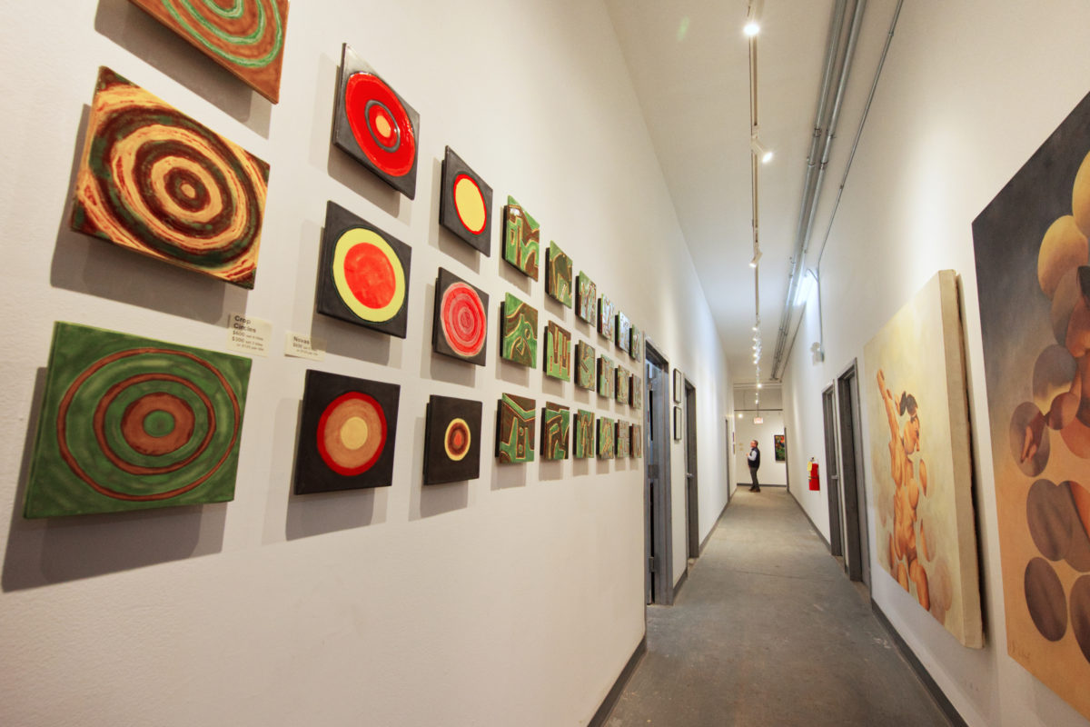 A long hallway with clean, white walls and warm, overhead lighting within an art studio setting. Shown hanging on both sides of the hall are a number of colorful ceramic tile artwork pieces and large, figurative oil paintings