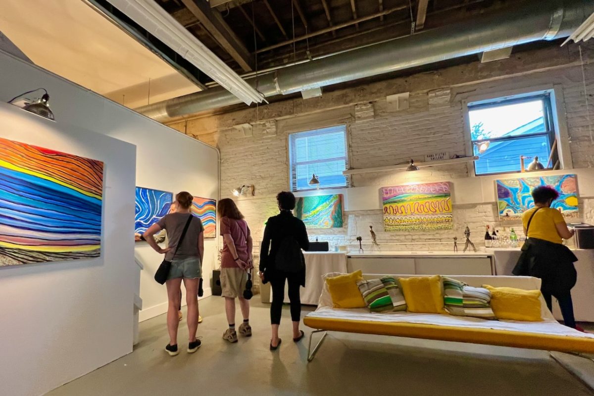 A view of three people from behind as they stand in an art studio looking at a row of colorful oil paintings hung on a wall. A window to the alley outside is behind them and a small, orange couch is visible in the right corner of the photo