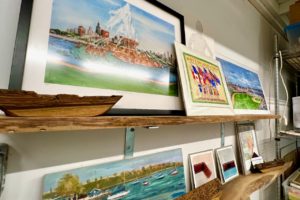 Two shelves of framed and unframed watercolor paintings of famous Chicago landmarks like the Buckingham fountain and the lake front, an overhead view of Wrigley field and a couple of smaller abstract artworks.