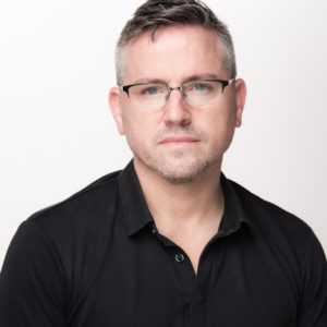 A man seated in a brightly lit space with a white background. He’s in his fifties but young-looking, with salt and pepper colored hair, a slight beard and rimmed eyeglasses. He’s wearing a black, collared shirt.