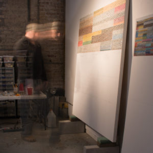 A man works in his studio. The man is seen on the left in a winter cap, dark green sweater and grey pants with a work table just in front of him. He's facing a large canvas almost as tall as his propped up on a white wall. On the canvas there are rows of old postage stamps he carefully arranges in rows by color to form an overall image of a pattern of colorful rectangles. The photo is taken with a time lapse effect to show the man appearing in a semi-transparent position and then another right next to it.