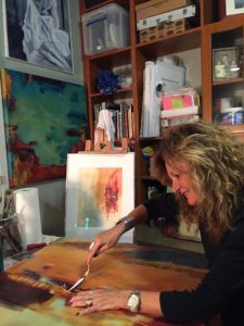 A woman of indeterminate middle age is seen working in her painting studio. She has shoulder-length, wavy, sandy-colored hair, a fair complexion and is wearing a black shirt and some jewelry. She’s shown using a palette knife to scrape along the edge of a canvas surface she’s painting on