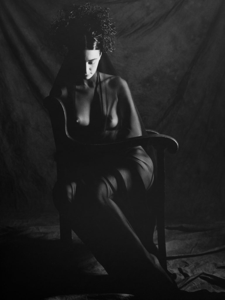 A women sits in a chair with her head down and eyes closed. She is covered by a sheer fabric gown. The light in the room is from a single point and stark, and the color scheme of the photo is black and white