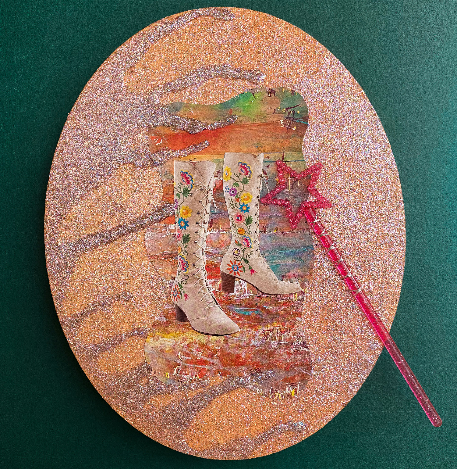 An oval-shaped piece of collage-art shown against a green background. The surface of the piece has an orange-pink color with a glitter finish. In the center there's a picture of a pair of women's boots with colorful floral patterns along the site and attached to the piece is a child's magic wand toy with a star-shaped tip.