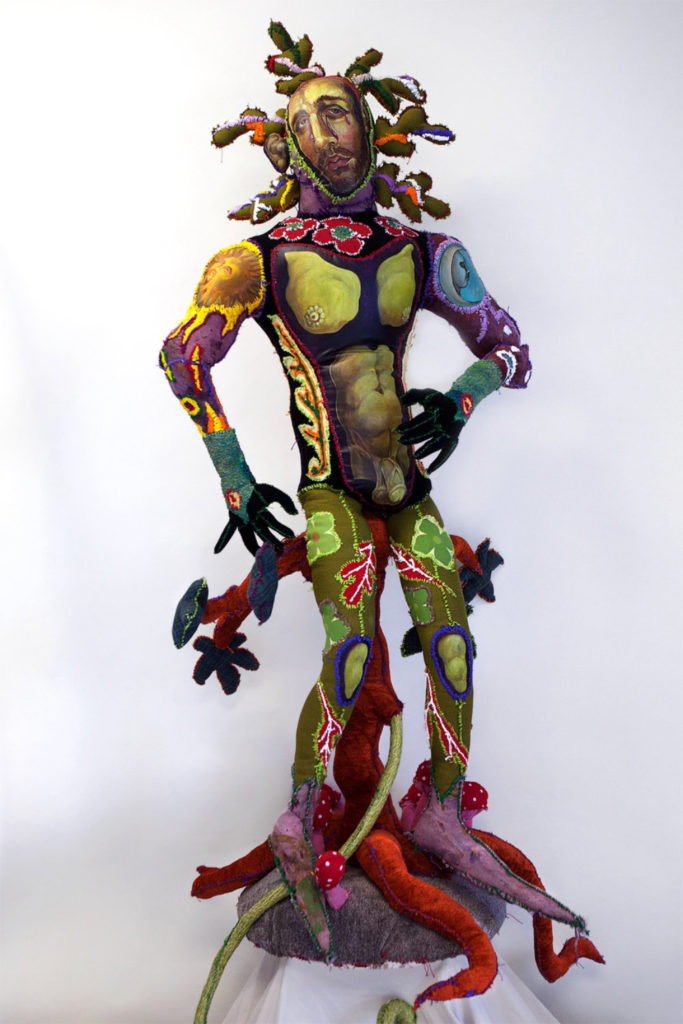 A sculpture of a nude male figure who's body is pieced together from different colored-and style of fabrics. The face is a painted Christ-like figure with cactus-like protuberences