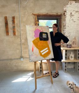 Artist Eva Pleuhs, a woman with medium-length brown hair, wearing eyeglasses and a black dress, is seen standing in a rustic art studio setting. The wall behind her is halfway covered with stucco, half exposed brick that's been painted white. Directly behind her is a window with a sunny day seen. And to her left some abstract clay sculptures sit on shelves improvised from cinderblock and flat boards. She's standing next to one of her paintings, which is about 3 feet tall by 2 feet wide and shows abstract gold, black and pink shapes with smaller brush strokes of varying color scattered throughout the surface. In the very bottom right of the photo, a small tan and white terrier-type dog is seen looking at the camera.