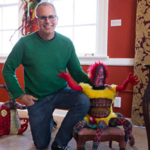 A middle-aged man in glasses, with jeans and a green sweater is kneeling next to a small, slightly menacing doll with numberous legs in plaid material, a yellow fur torso with teeth in the middle, red outstretched arms, a painted-on red face and frizzy red hair. They're both seen in a red room that appears to be part of an art exhibit