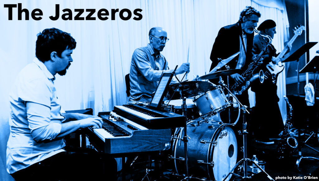 A jazz band putting on a performance. A man with dark hair, trim beard and wearing a white shirt sits playing a jazz organ in the foreground. A thin, middle-age man with trim beard and glasses is playing a drum kit next to him, behind him is a middle-age man with a dark sport jacket on playing tenor sax, and all the way to the right (in the rear from the photo perspective) another man of middle age plays a black and white electric bass guitar.