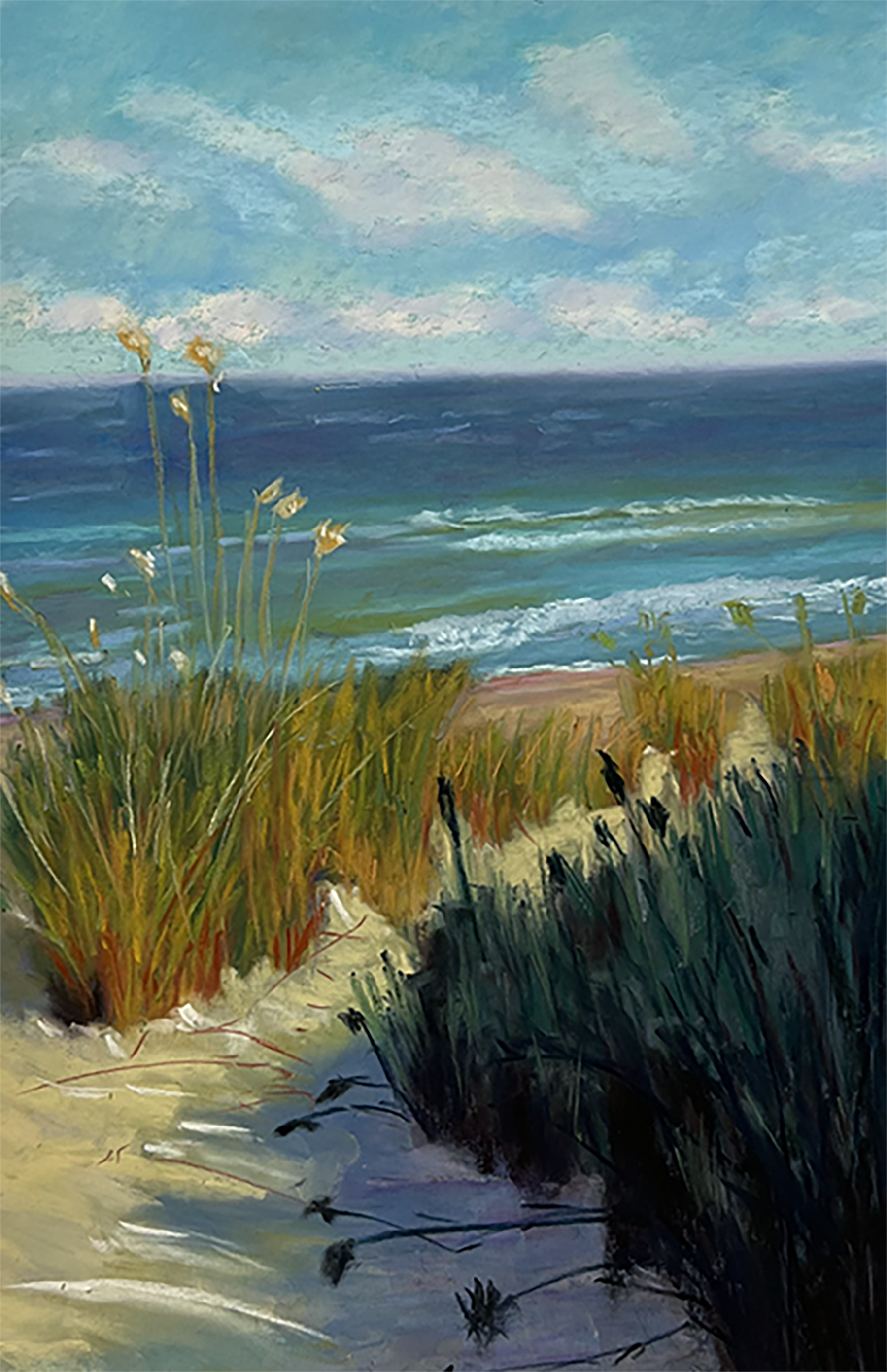 A sandy beach with brush growing in the foreground is shown on a bright day with clouds and rolling waves from a deep blue sea in the background.