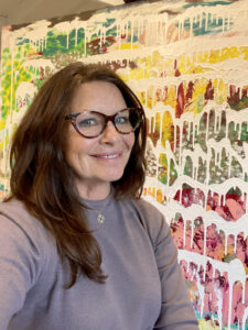 A woman with a light Mediterranean complexion and medium brown hair is seen in a lavender shirt and brown-rimmed glasses is seen standing in front of an abstract painting composed of colorful splatters of red, green, yellow and pink paint with rows of dripping white paint running across