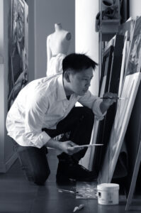 A man in a white shirt and jeans with short, dark hair is seen in a studio on one knee painting on a canvas that's on the floor propped up against a wall. The photo is in black and white.