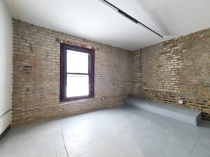 A studio space with a window is brightly-lit by natural daylight coming in. Three brick walls have outlets mounted along them.