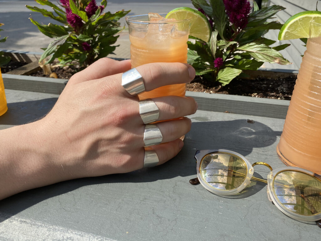 A hand seen up close is on a table holding an orange, iced beverage on a sunny afternoon. A large, metallic ring is seen on each finger.