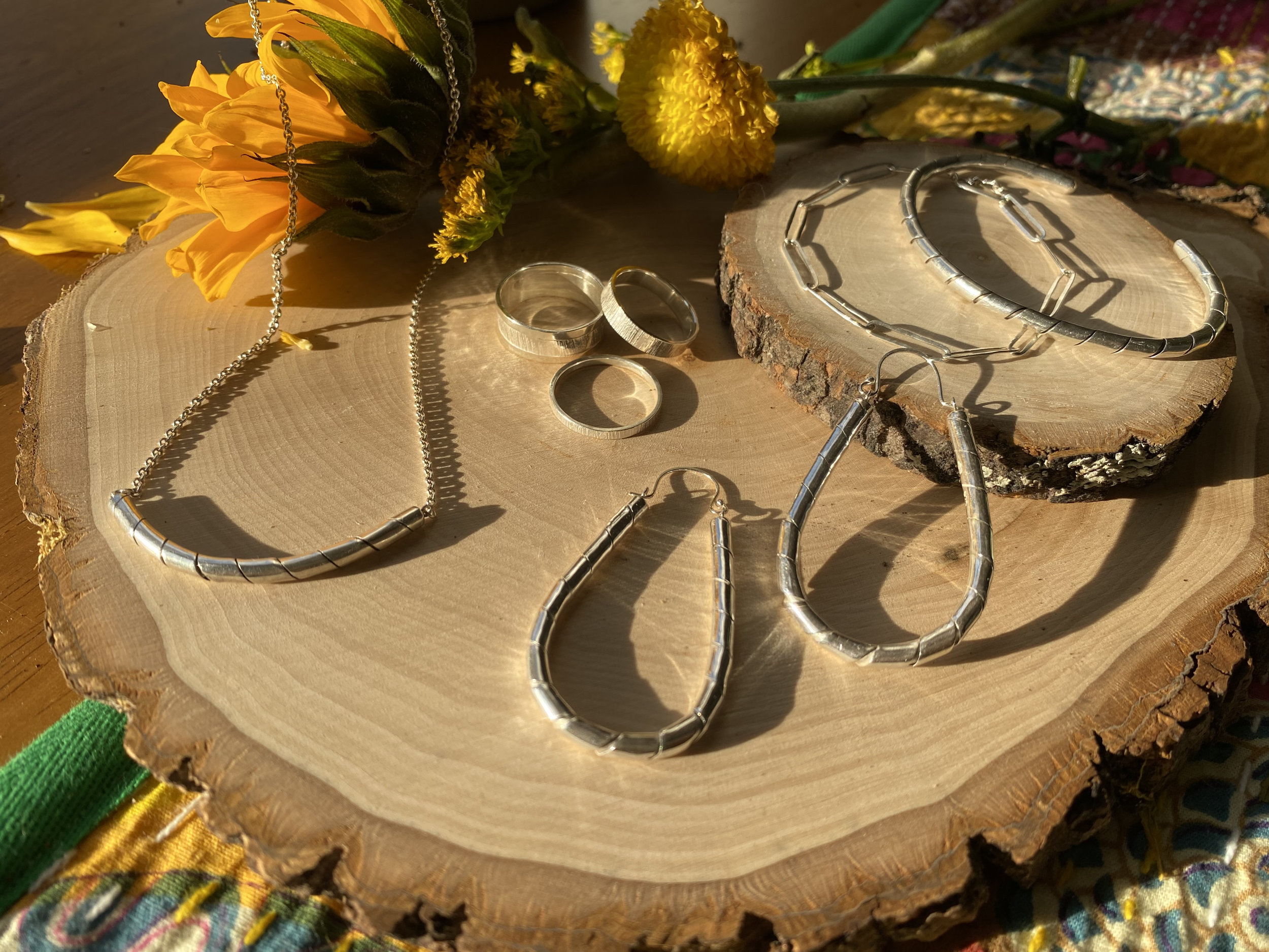 An array of rings, large earrings, a necklace and a bracelet are on a section of a wood with some sunflowers in warm, afternoon sunlight