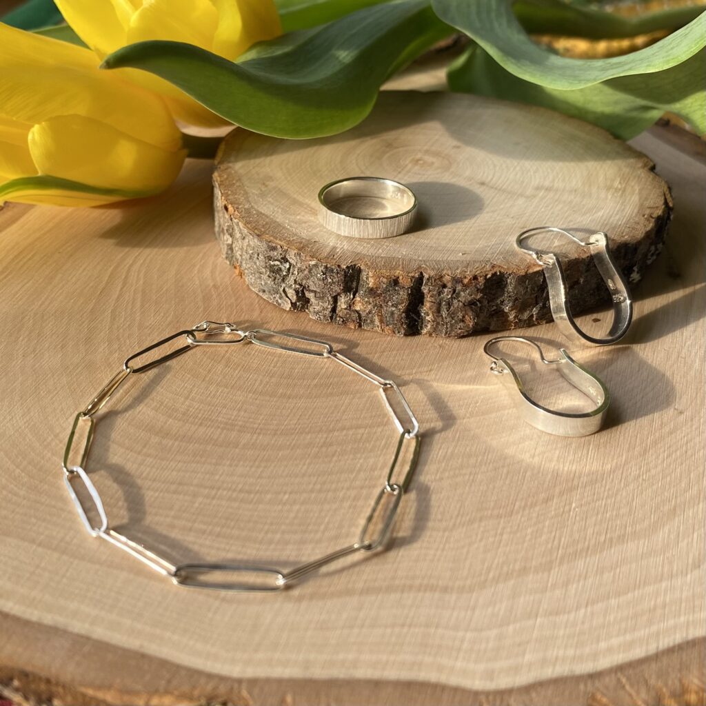 Two earrings that have a curved shape, a metal ring and a bracelet composed of simple metal links are laid out on two sections of a tree trunk with a tulip laying in the background in warm, afternoon light.