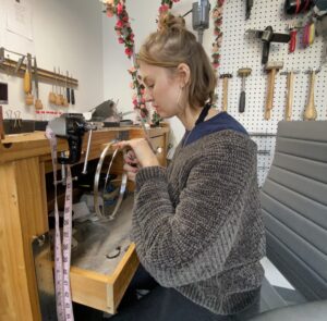 A woman with neck-length sandy-blonde hair, wearing large earrings, a brown sweater and blue shirt is seen in side profile in her studio at a work bench with various tools for metal-working. Behind here is a column of flowers going up to the ceiling alongside a pegboard with more metalworking hammers, awls and saws.