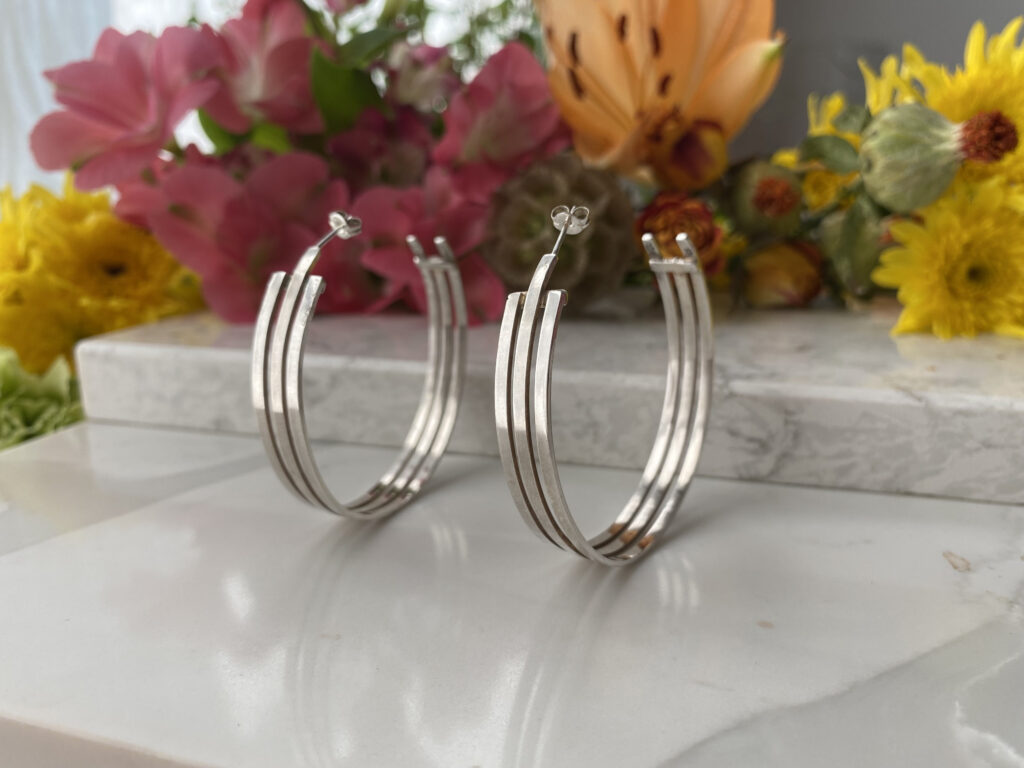 Two earrings composed of single, curving pieces of metal are seen on a metal countertop with an assortment of flowers of different colors in the background.