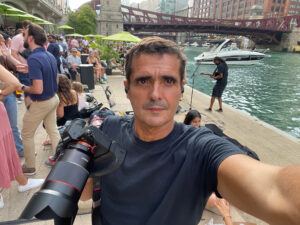 A man in his mid-forties with short, brown hair and a tanned complexion and wearing a black t-shirt is holding a large digital camera with long lens attached. Behind him is the Chicago River on a very summery day with a. large crowd watching a guitarist perform and several boats in the water nearby.