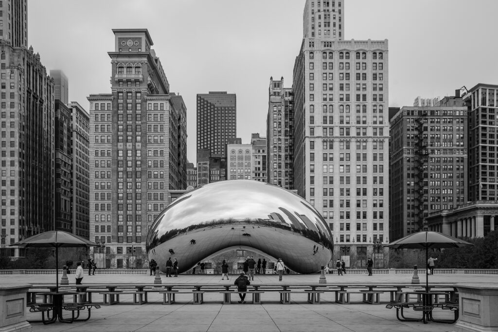The Chicago sculpture "Cloudgate" – also known as "The Bean" taken from straight ahead in black and white. The shiny metallic sculpture is seen against a number of large skyscrapers. Several people are seen wandering around both under the sculpture and in the foreground