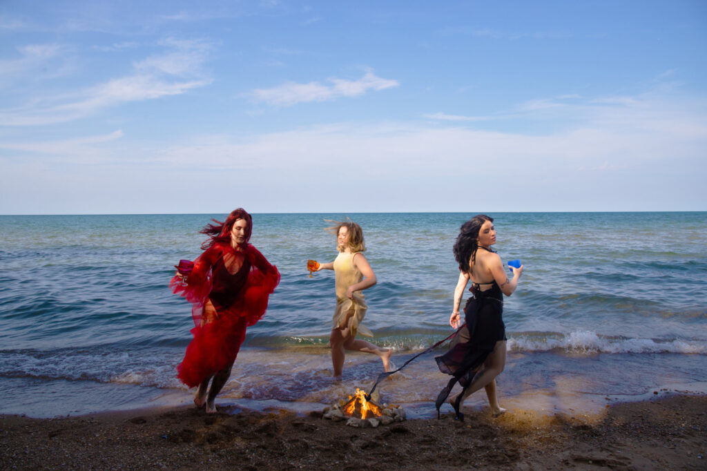 Three women roughly in their twenties – with red hair and wearing a sheer red dress, another with dark blonde hair and a wearing tan, light fabric dress, and a third with dark hair and a flowing black dress – dance around a fire along the shore of an open sea. Each is holding a chalice as she dances
