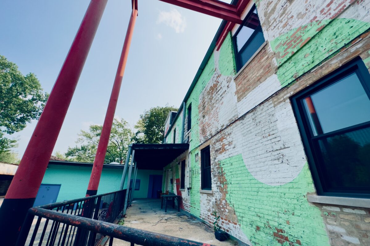 The loading dock of an old brick building covered in light green and white paint spelling out the word "Arts" on a sunny summer afternoon. Some industrial elements are seen attached to the building, including a set of large steel beams protruding out that have been painted red.