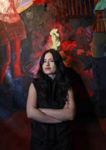 A young woman with dark, flowing hair, a fair complexion and wearing a black, sleeveless vest and suit, is seen standing with arms crossed looking directly at the viewer while in a darkened, red and black room with painted imagery of a fire and women dancing on wall behind her.