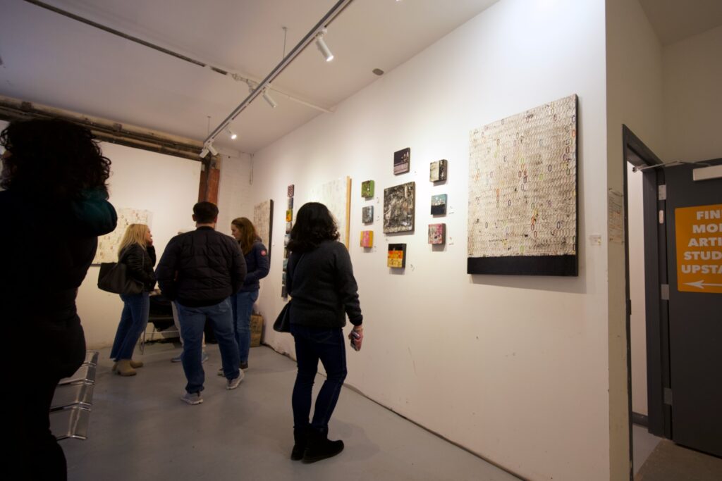 Several people seen standing in an art gallery setting viewing a series of paintings with (indiscernible) modernist subject matter on a wall in a clean, white, minimalist space