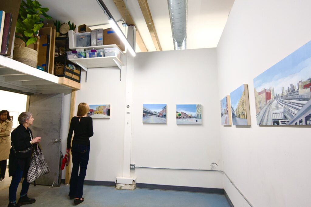 A younger woman and older woman standing in a clean, white-walled art space viewing a series of small cityscape paintings that take up a pair of walls in a single row.