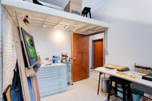 An occupied art space with warm lighting, a work table with materials, a painting on an easel and a filing drawer. The space is bright with clean, white walls
