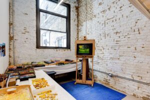 An artist studio with tall white-washed brick walls, an easel with a small painting and an open window letting bright daylight into the space