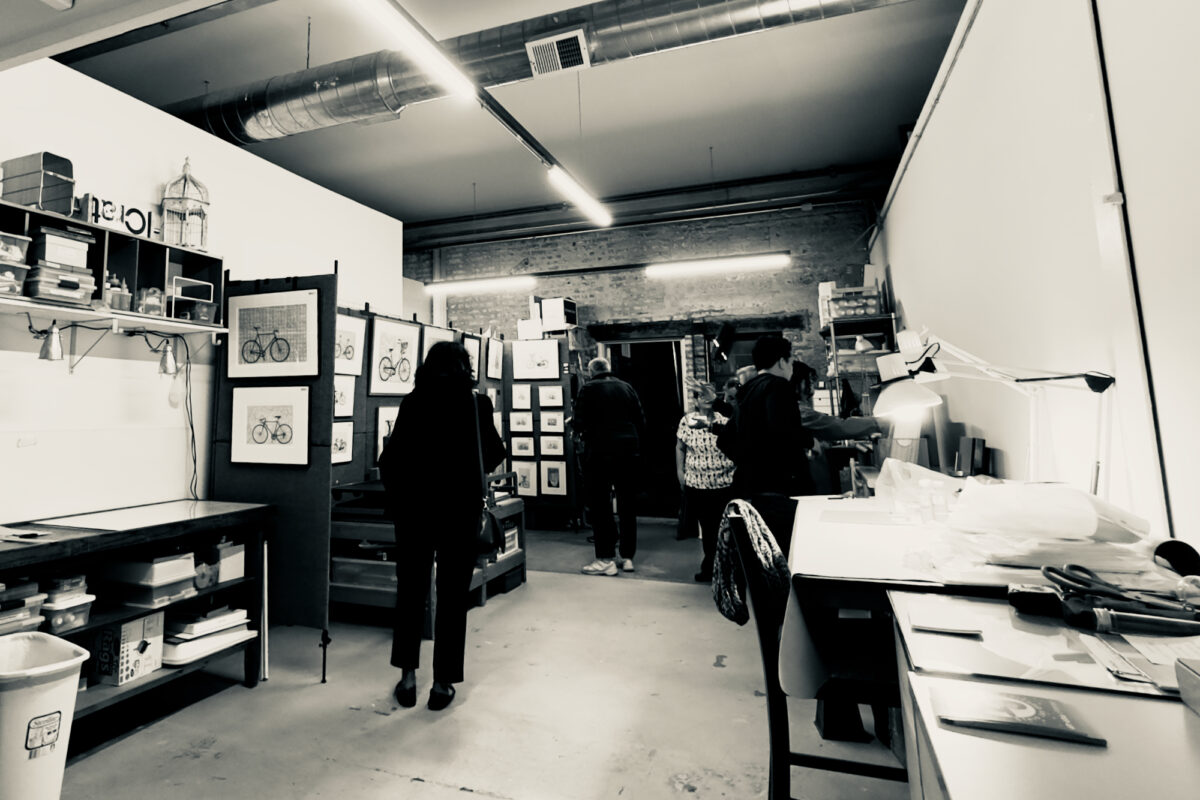 Several men and women view fine art prints in a brightly-lit work studio. Work desks and tables with art materials and tools are seen throughout the space along with some display walls where framed prints are hung for visitors to look at