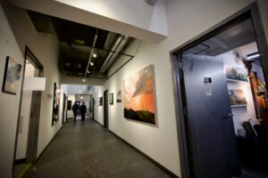 A long, brightly-lit hallway with white walls is adorned with various types of colorful paintings. A studio door is open with artwork and work materials visible inside. At the end of the hall, two people can be seen looking at the artwork.